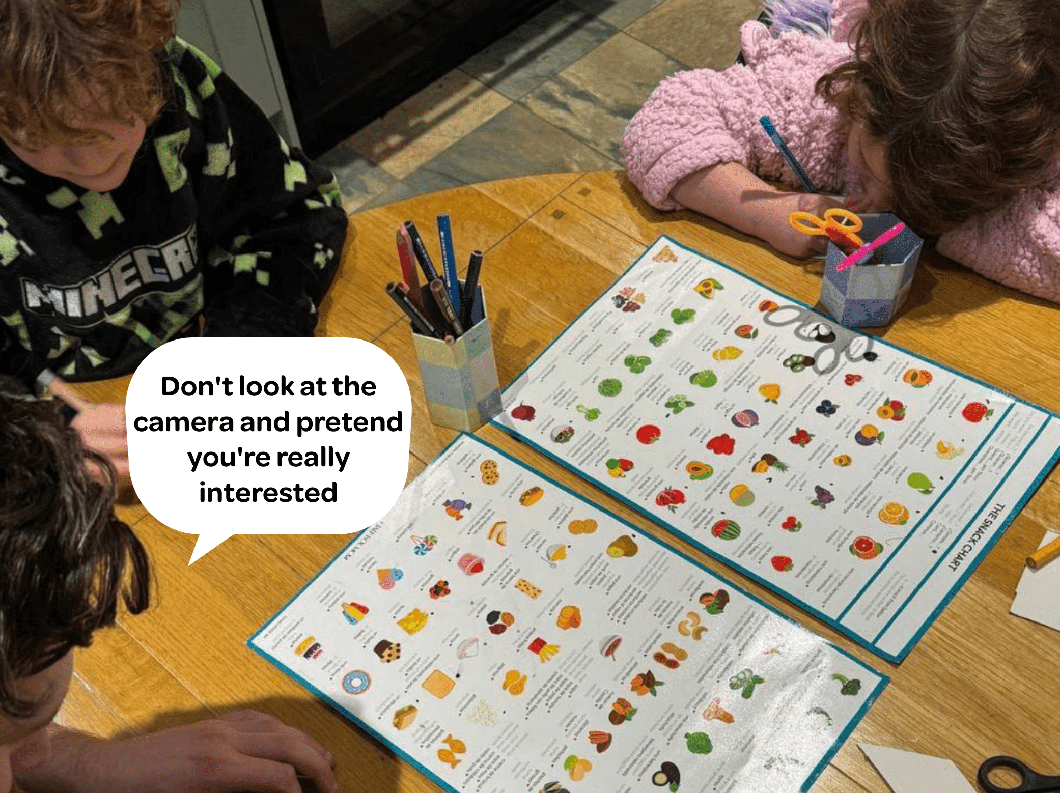 Two kids doing a homeschooling activity at a table, looking at a colorful laminated chart