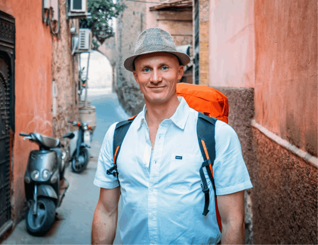 Matt Karsten of Expert Vagabond smiling in a hat with a backpack in a colorful alley — top travel blog for adventure, photography, and long-term travel tips.