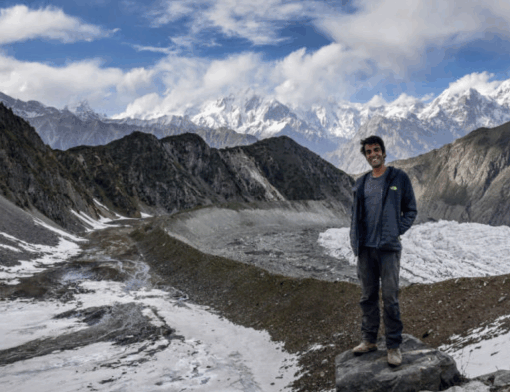 Joao from Against the Compass stands on a mountain trail surrounded by snow-capped peaks in Pakistan.